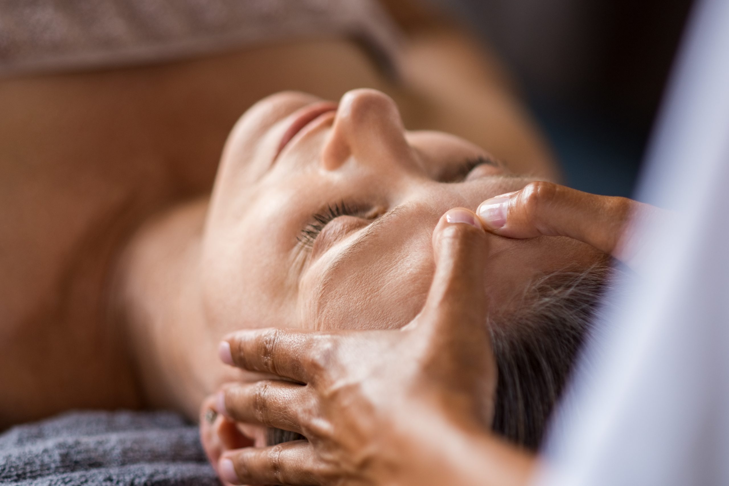 Closeup face of mature woman having facial massage at spa. Senior woman lying with closed eyes at spa while a massage therapist doing anti-aging treatment. Masseur doing head massage at wellness center.